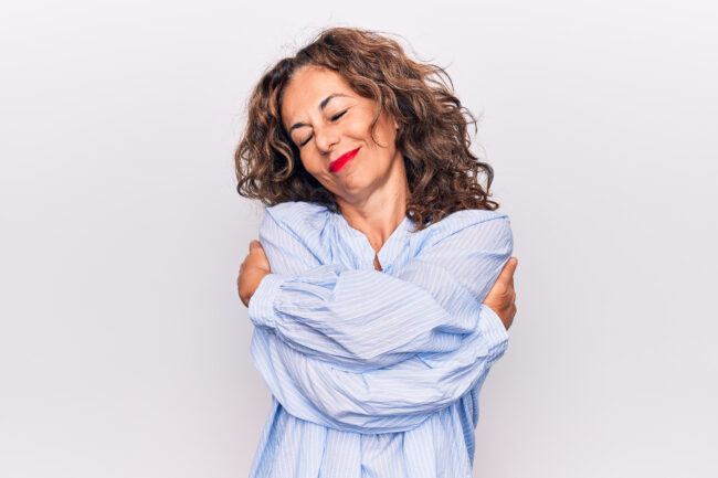 Middle age beautiful brunette woman wearing striped shirt standing over white background hugging oneself happy and positive, smiling confident. Self love and self care