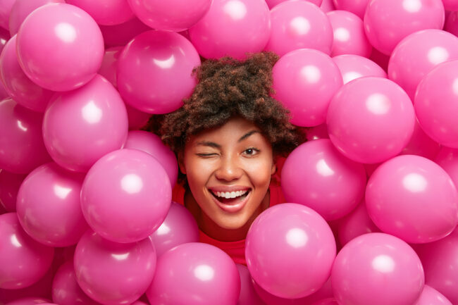 Optimistic birthday girl has fun and winks eye smiles joyfully poses against many pink air balloons spends time on party and enjoys happy celebration came to congratulate best friend with anniversary